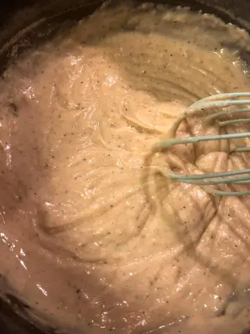 Sauce for macaroni and cheese in a saucepan with a blue whisk.