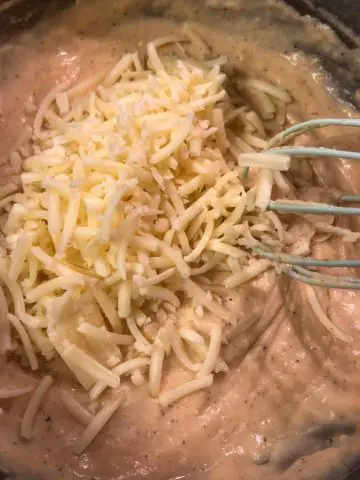 Sauce for macaroni and cheese in a saucepan with shredded white cheddar and a blue whisk.