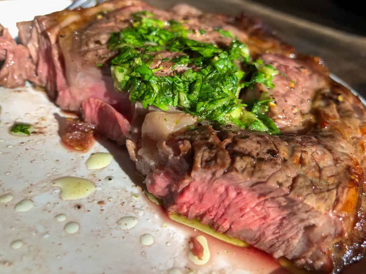 Ribeye steak which has been cut to show it is cooked medium rare with juices and olive oil surrounding the steak and topped with a cilantro and garlic sauce on a white plate.