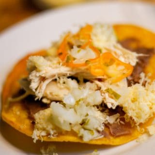 Belizean garnaches which is a corn tostada topped with refried beans, chicken, shredded cheese and relish on a white plate with cheese in the background.