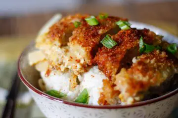 Breaded pork chop pieces on top of rice in a bowl and green onions garnishing the top of the pork chop pieces.