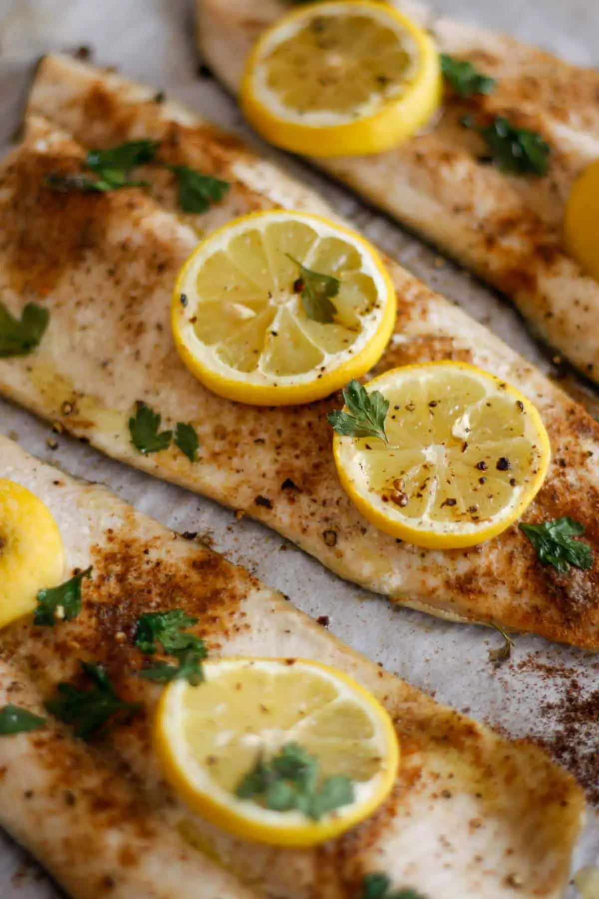 Baked Rainbow Trout on parchment paper sprinkled with Old Bay Seasoning topped with slices of lemon and Italian parsley.