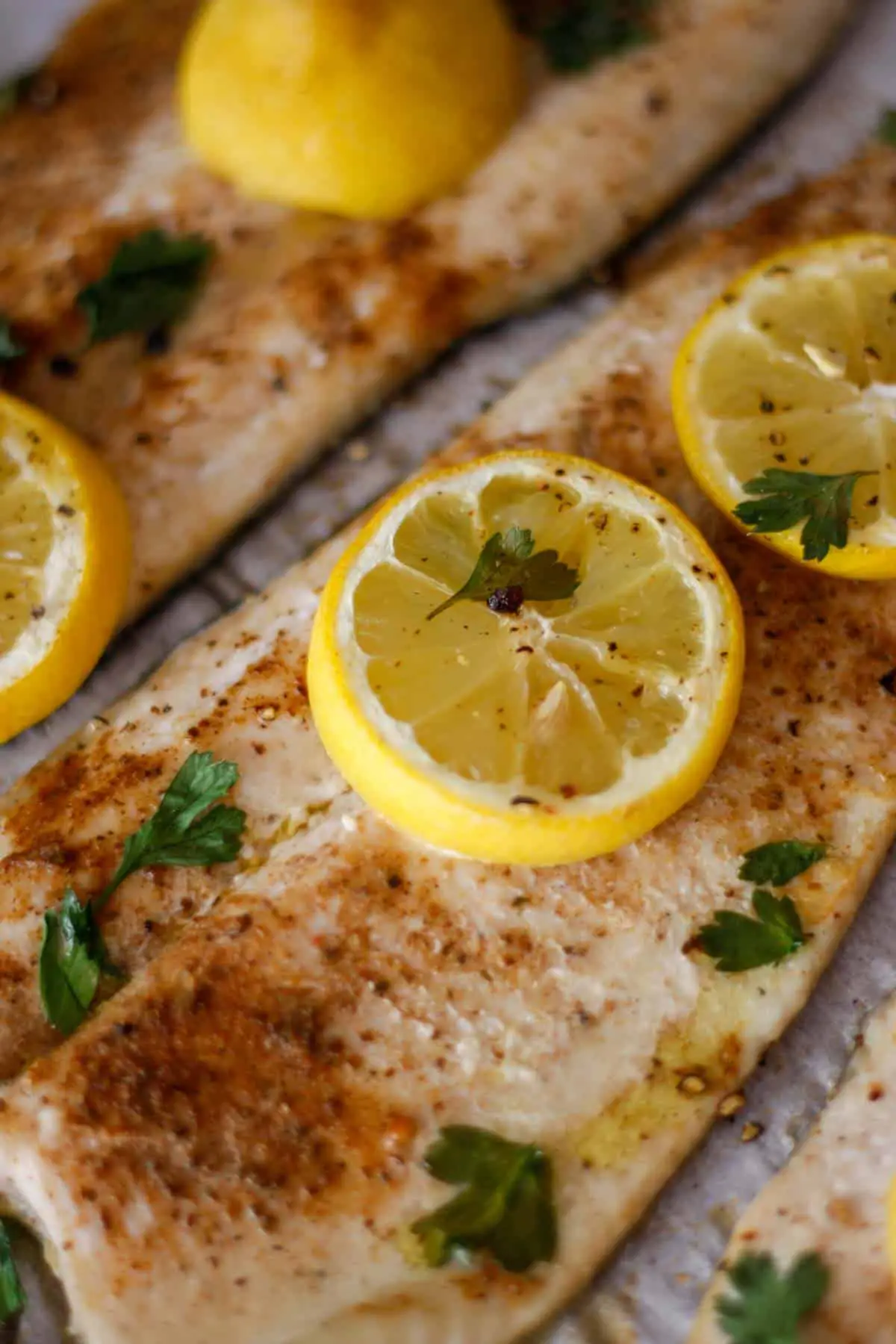 Baked Rainbow Trout on parchment paper sprinkled with Old Bay seasoning topped with slices of lemon and Italian parsley.