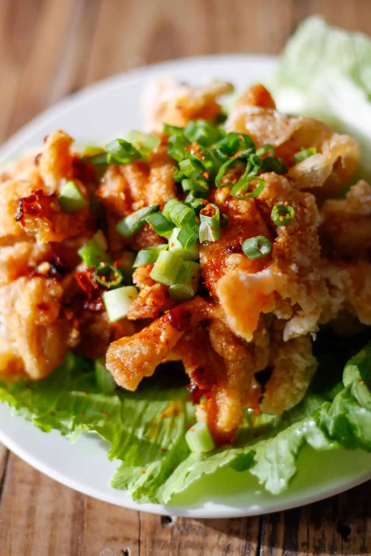 Pieces of fried chicken atop iceberg lettuce drizzled with a soy based sauce and topped with sliced green onions on a white plate.