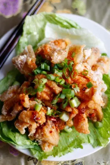 Pieces of fried chicken atop iceberg lettuce drizzled with a soy based sauce and topped with sliced green onions with chopsticks on the left hand side.