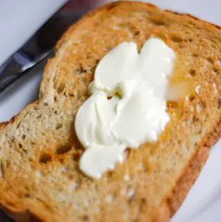 Piece of buttered rye bread toast on a white plate with a knife on the left hand side.