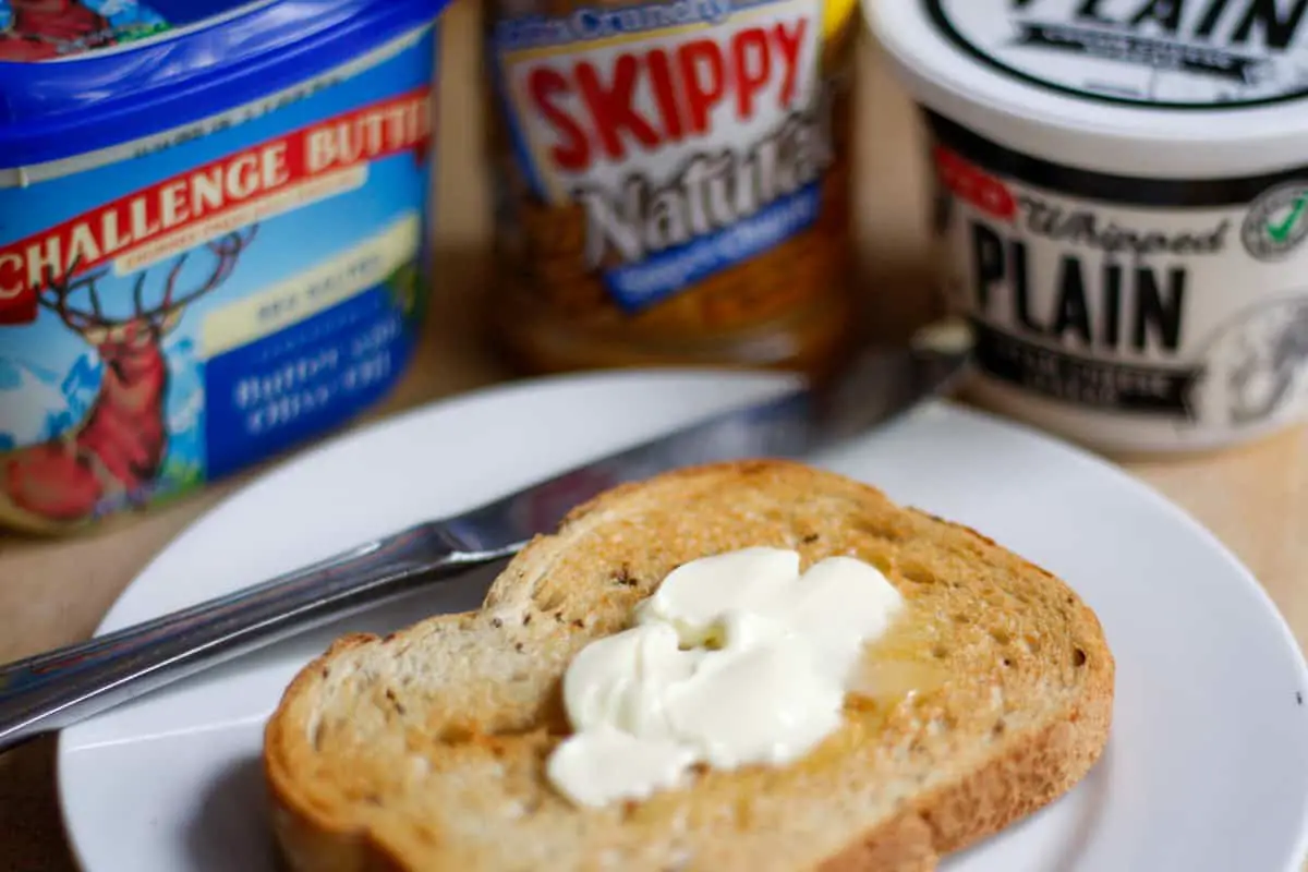 Piece of buttered rye bread toast on a white plate with a knife on the left hand side and cartons of Challenge butter, Skippy Peanut Butter, and Whipped Plain Cream Cheese Spread in the background.