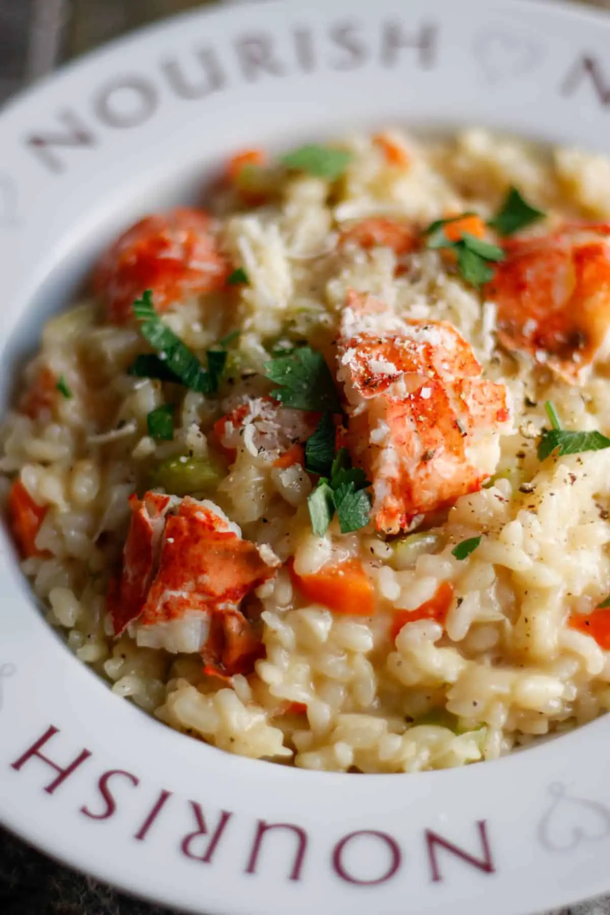 Lobster risotto garnished with parsley and Parmigiano Reggiano in a white bowl with the word Nourish on it and hearts.