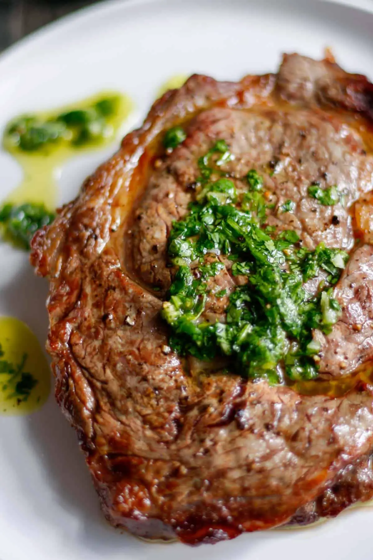A cooked ribeye steak on a white plate which is topped with a cilantro garlic sauce and the sauce is also drizzled on the plate next to the steak.