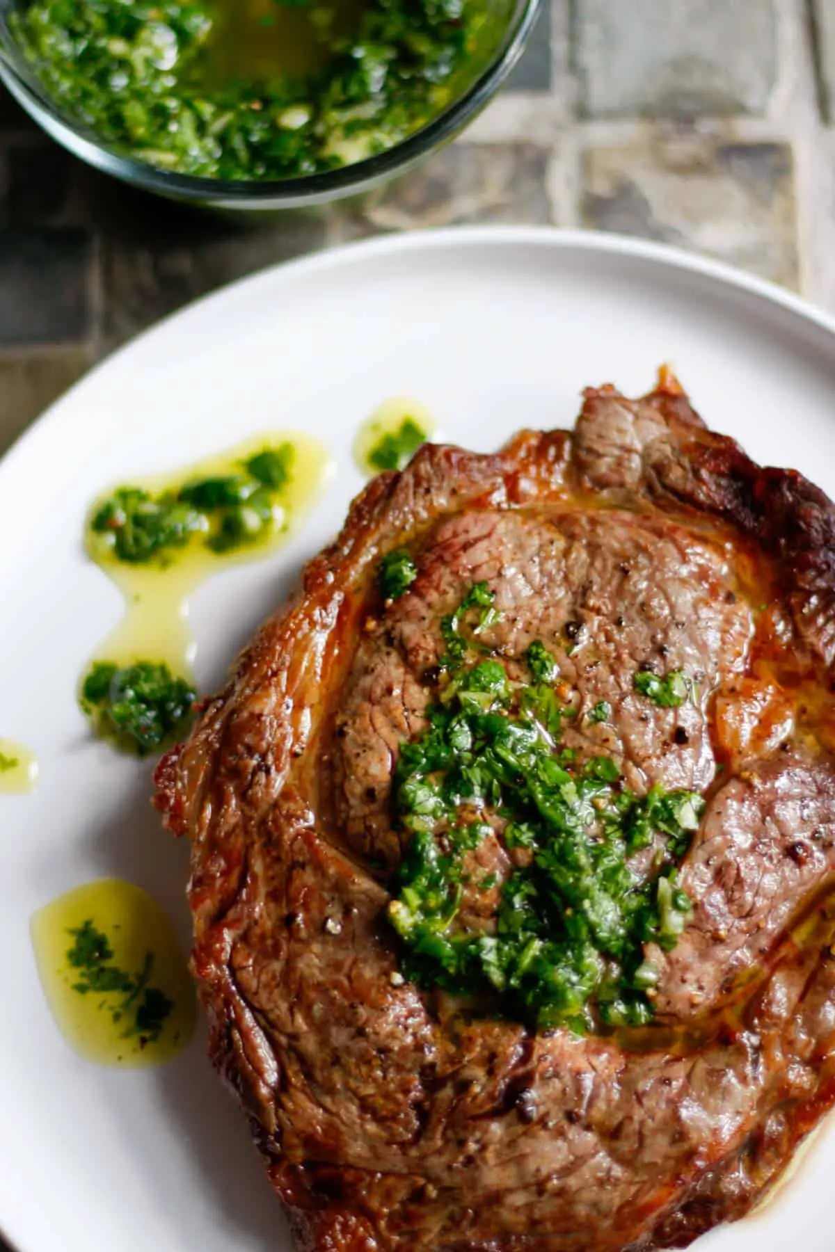 A cooked ribeye steak on a white plate which is topped with a cilantro garlic sauce and the sauce is also drizzled on the plate next to the steak with a glass bowl containing the cilantro garlic sauce in the background.