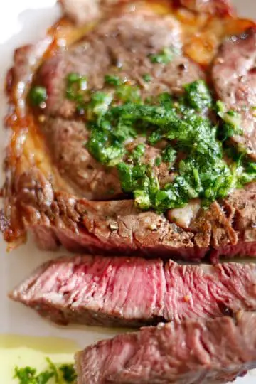 Ribeye steak which has been cut to show it is cooked medium rare with juices and olive oil surrounding the steak and topped with a cilantro and garlic sauce on a white plate.