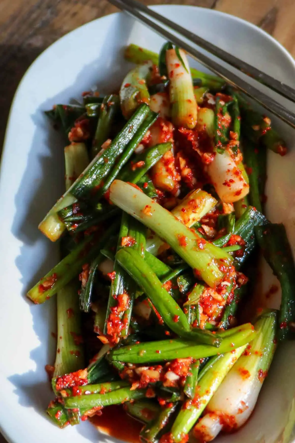Sliced green onions in a red pepper kimchi sauce placed in a white dish with silver chopsticks resting on the side.