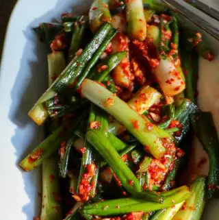 Sliced green onions in a red pepper kimchi sauce placed in a white dish with silver chopsticks resting on the side.