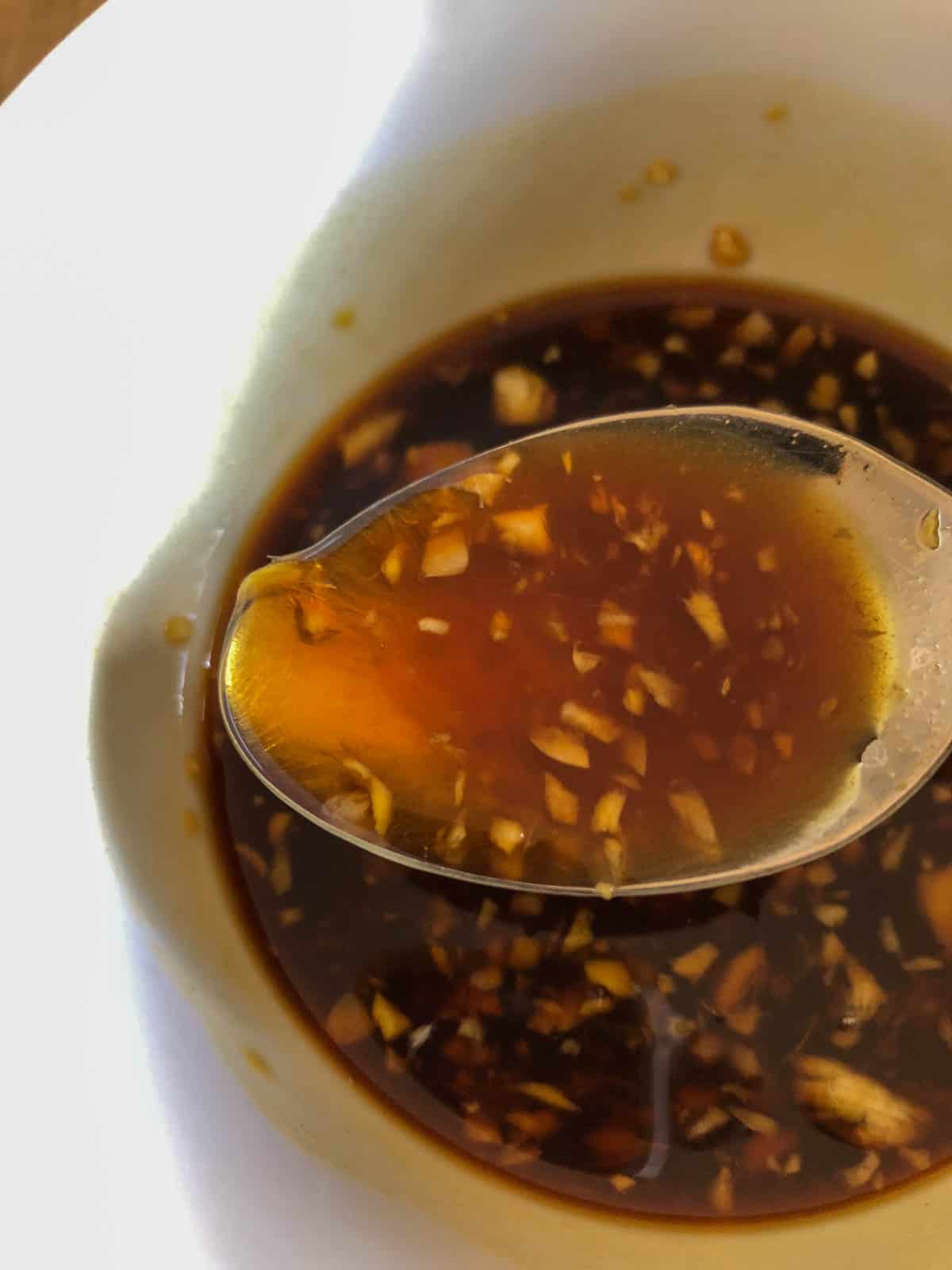 A soy sauce based sauce with pieces of ginger and garlic in a white bowl with a spoon containing some of the sauce in the foreground.