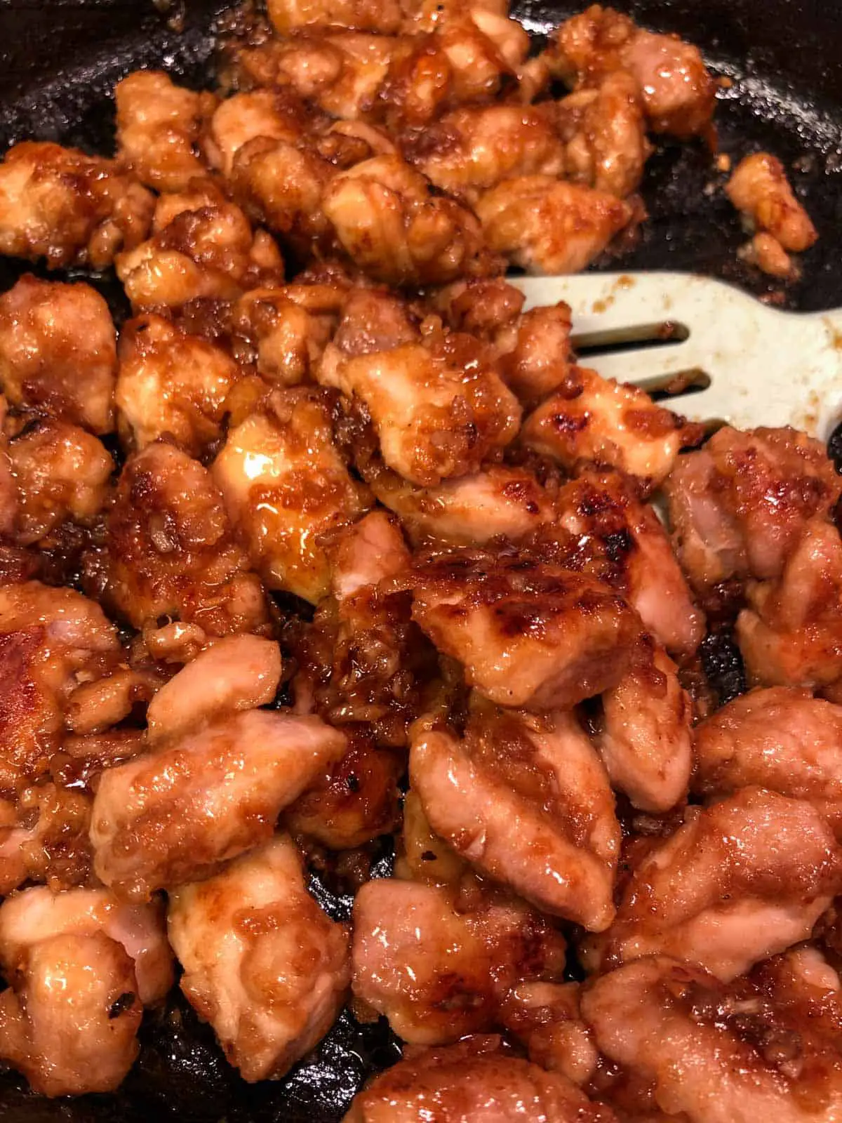Chicken pieces coated with a sticky sauce in a cast iron pan with a blue utensil.