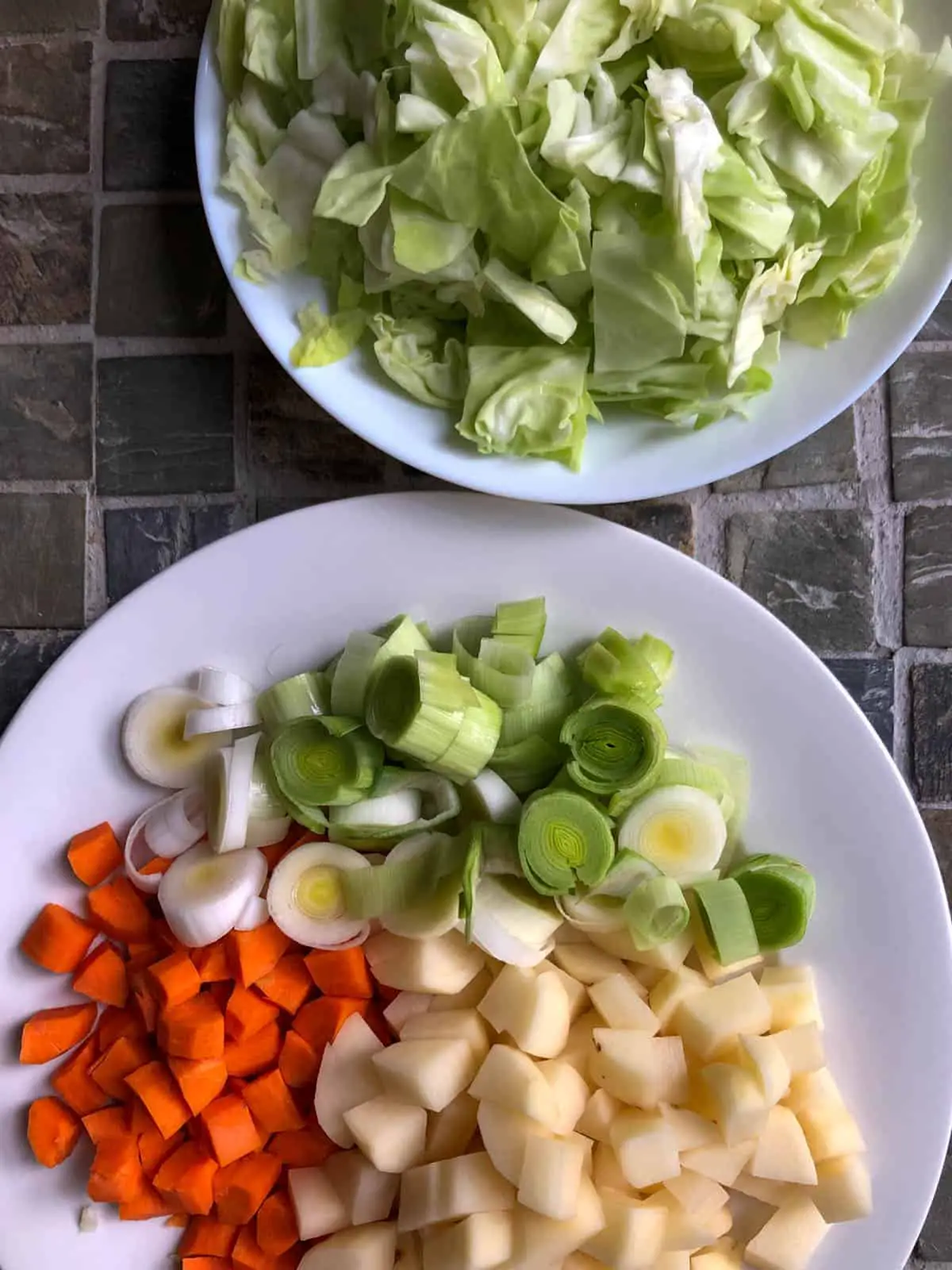White bowl filled with cut up pieces of cabbage and another white bowl filled with cut up carrots, cut up leek, and cut up potatoes.