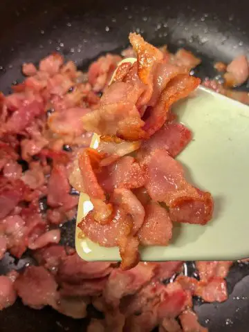 Diced cooked crispy bacon with bacon fat and a blue spatula holding some of the cooked bacon in the foreground.