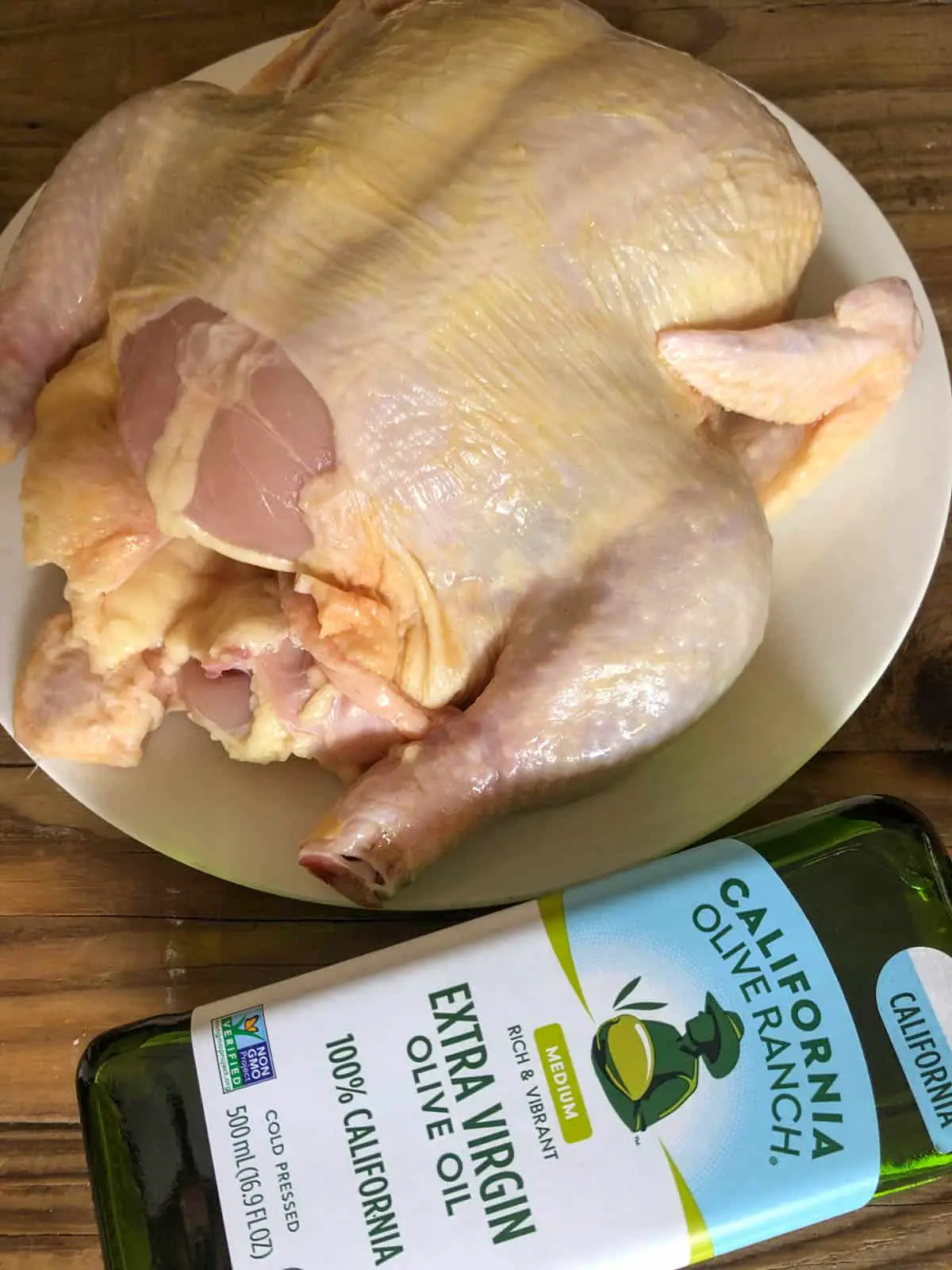 An uncooked whole chicken on a white plate with a bottle of extra virgin olive oil next to it.