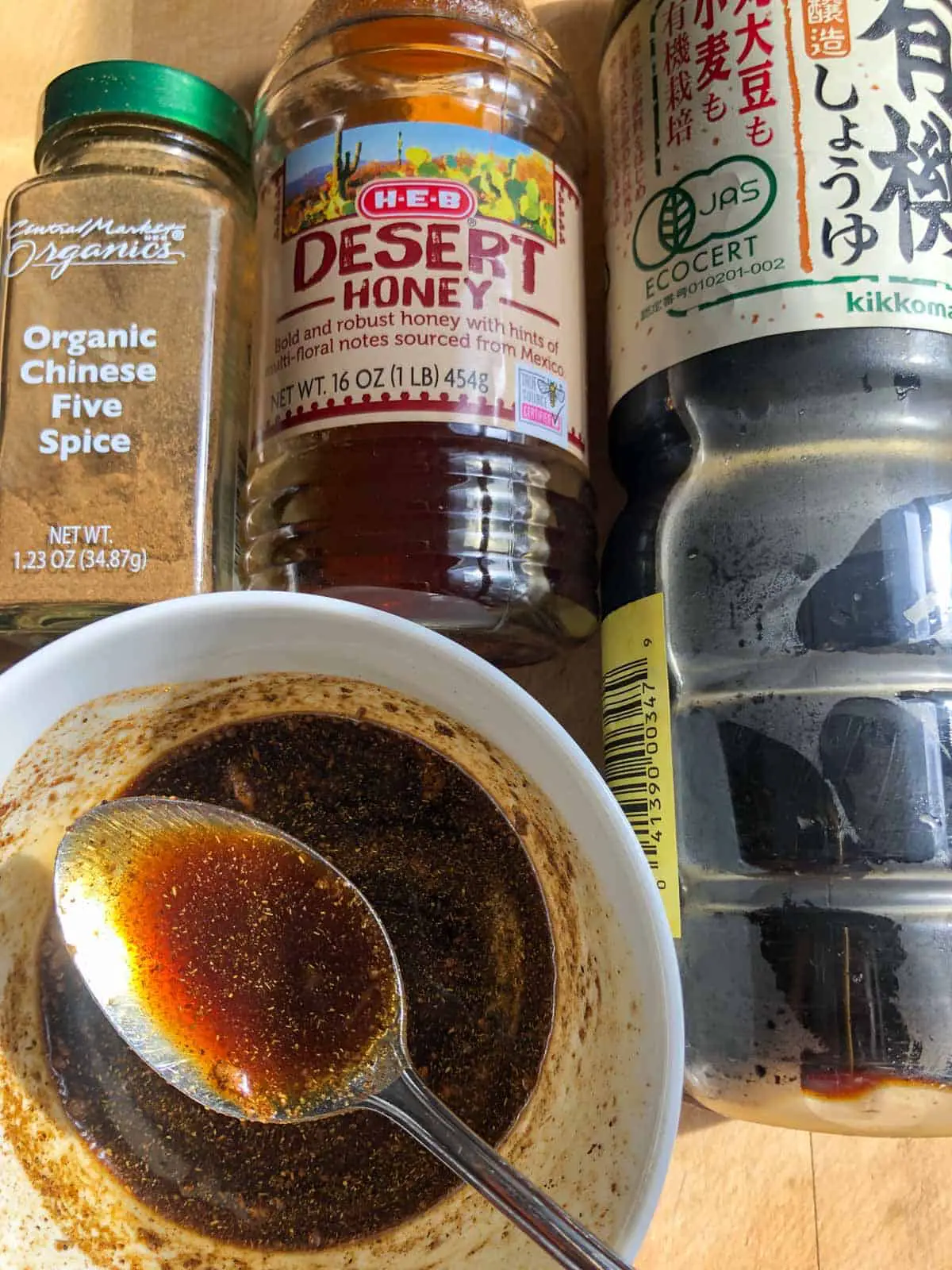 Bottle of Chinese 5 spice, bottle of honey, bottle of soy sauce, and a white bowl filled with a mixture of these ingredients and a spoon over the bowl containing this sauce.