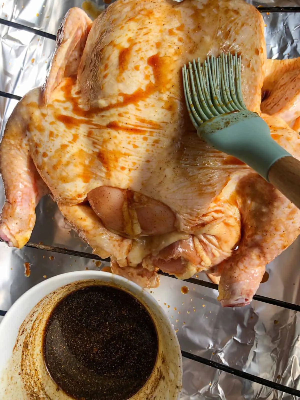 An uncooked whole chicken on a roasting rack with a blue baster basting the chicken with a soy sauce based sauce with Chinese 5 spice and honey and a white bowl containing the sauce next to the chicken.