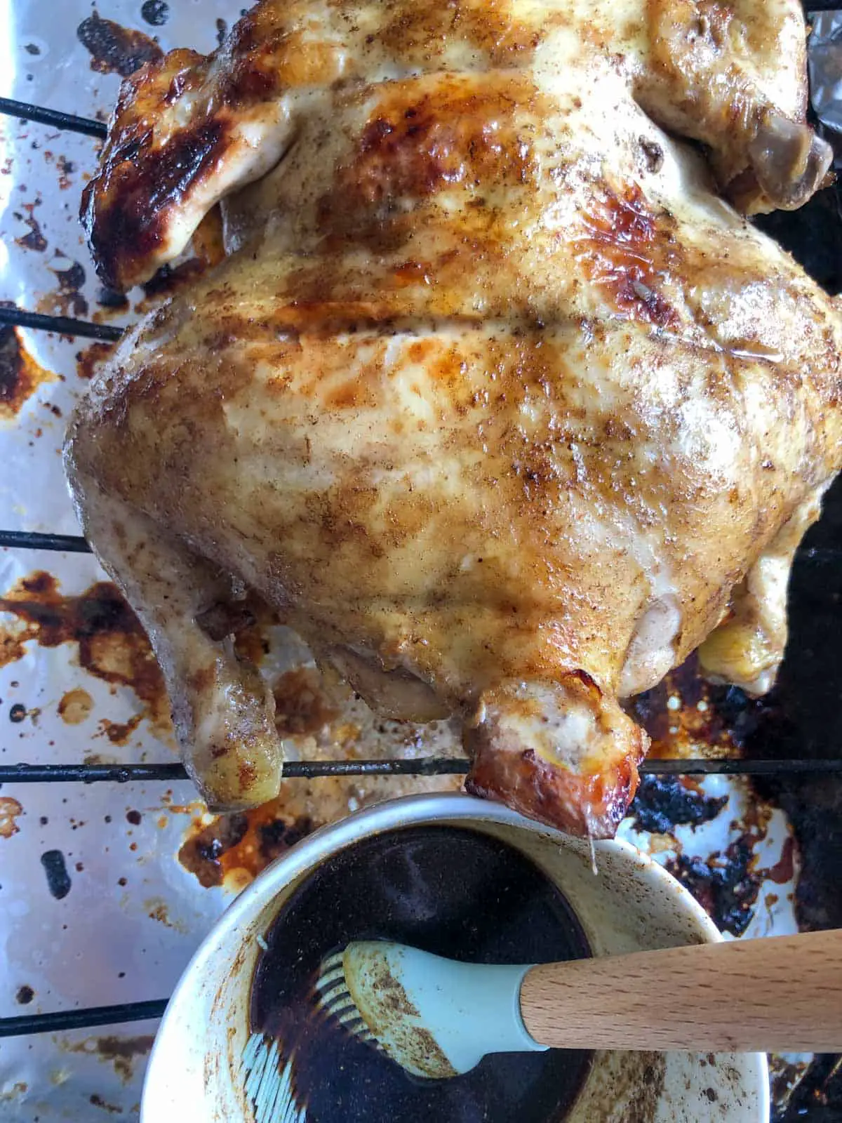 Whole chicken that has been browned in the oven on top of a roasting rack with a white bowl filled with soy sauce, honey, and Chinese 5 Spice with a blue baster next to the chicken.
