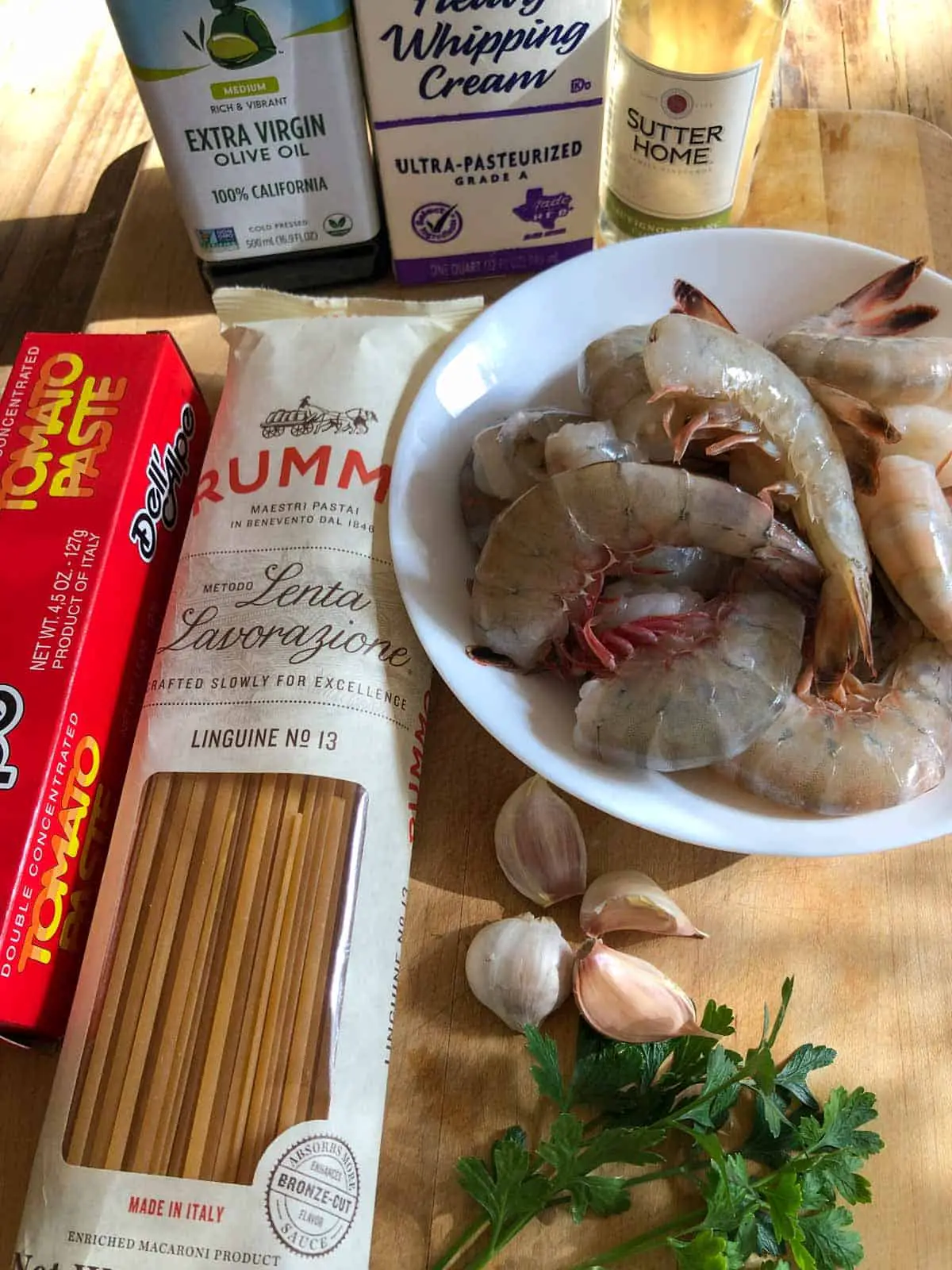 Shrimp with shell on in a white bowl, bottle of white wine and extra virgin olive oil, carton of heavy whipping cream, box of tomato paste, package of linguine, 4 garlic cloves and Italian parsley