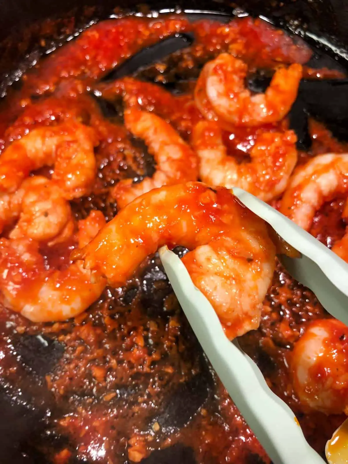 Red pieces of shrimp that have been coated with tomato paste with blue tongs holding one piece of shrimp.