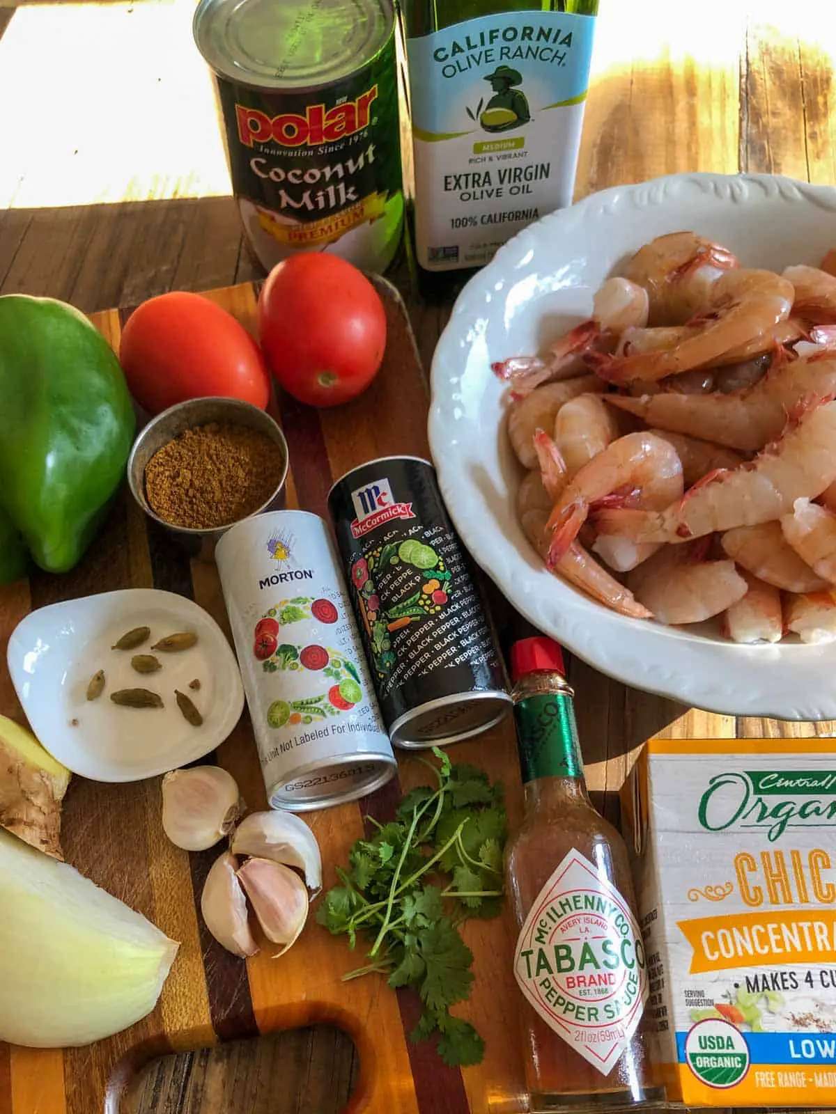 Can of coconut milk, bottle of olive oil, white bowl filled with uncooked shrimp, box of chicken broth, bottle of Tabasco sauce, cilantro, 4 cloves of garlic, salt and pepper, onion, ginger, cardamom pods on a white dish, green bell pepper, garam masala in a silver dish, 2 roma tomatoes.