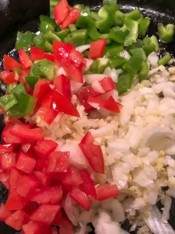Diced green bell pepper, diced onions and diced tomatoes in a cast iron pan.