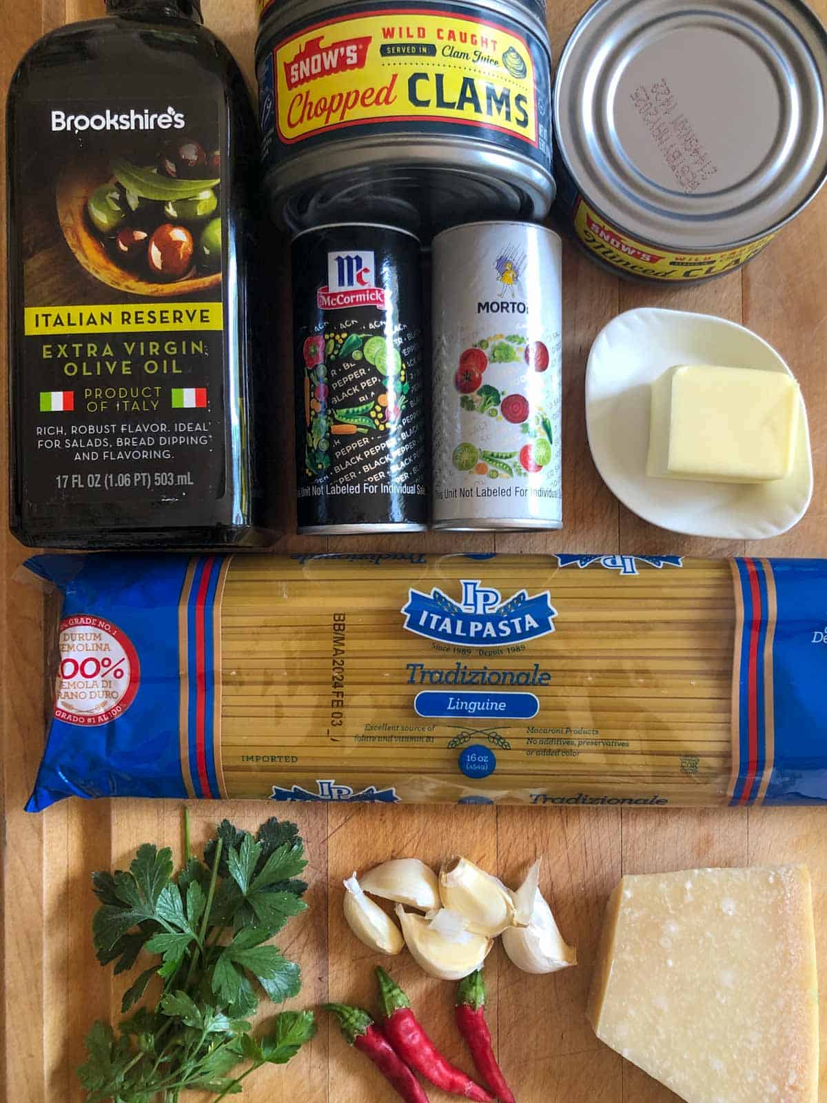 Bottle of olive oil, cans of chopped clams, salt and pepper shakers, butter on a white dish, package of linguine, Italian parsley, thai chilies, garlic, and a block of Parmesan cheese