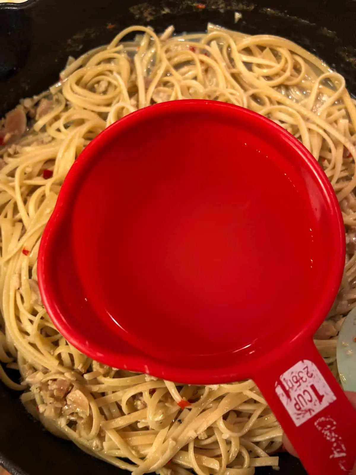 Linguine in the background in a pot with a red measuring cup in the foreground that contains pasta water.