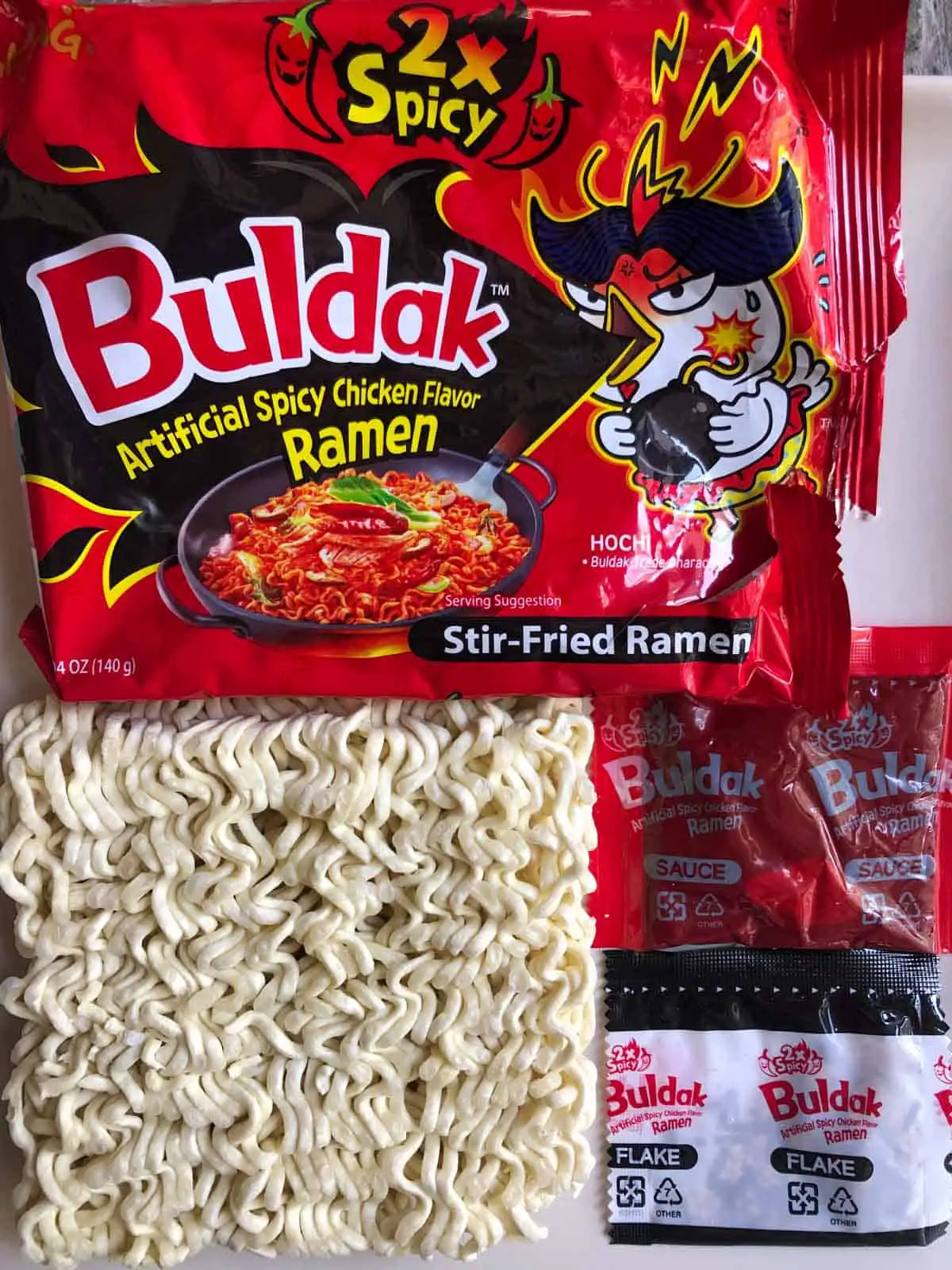 Packaging for Samyang Buldak 2x Spicy Ramen, the noodles that come in the packet, a packet of spicy sauce, and a packet of flake.