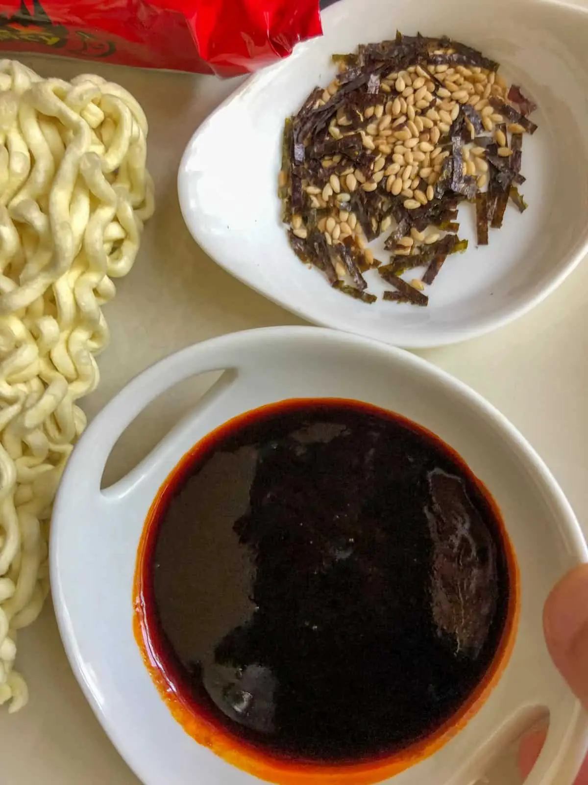 Some noodles, red packaging, seaweed and sesame seeds in a white dish, and a spicy sauce in a white bowl.
