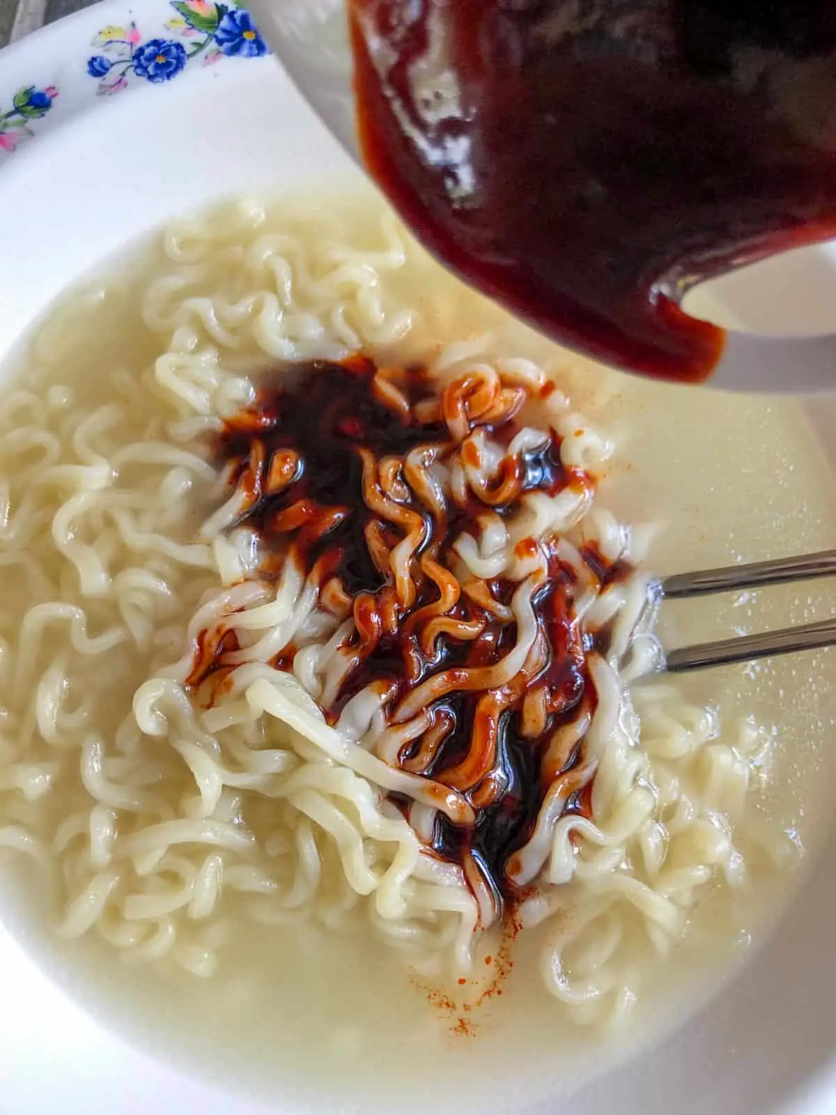 Noodles in starchy water in a white bowl patterned with blue flowers with spicy red sauce that has been added to the top of the noodles and a set of silver chopsticks.