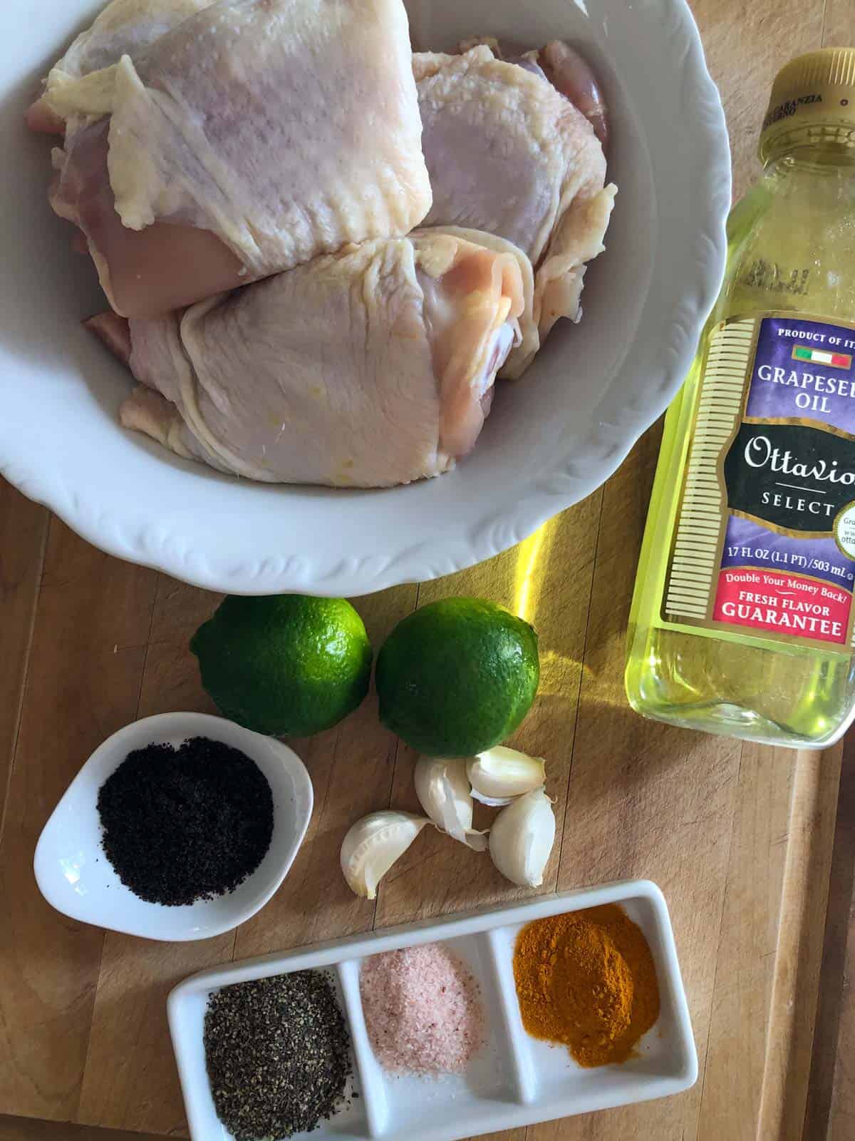 Chicken thighs with skin in a white bowl, a bottle of grapeseed oil, 2 limes, 4 cloves of garlic, sumac in a white dish, salt, pepper, and turmeric in a white dish.