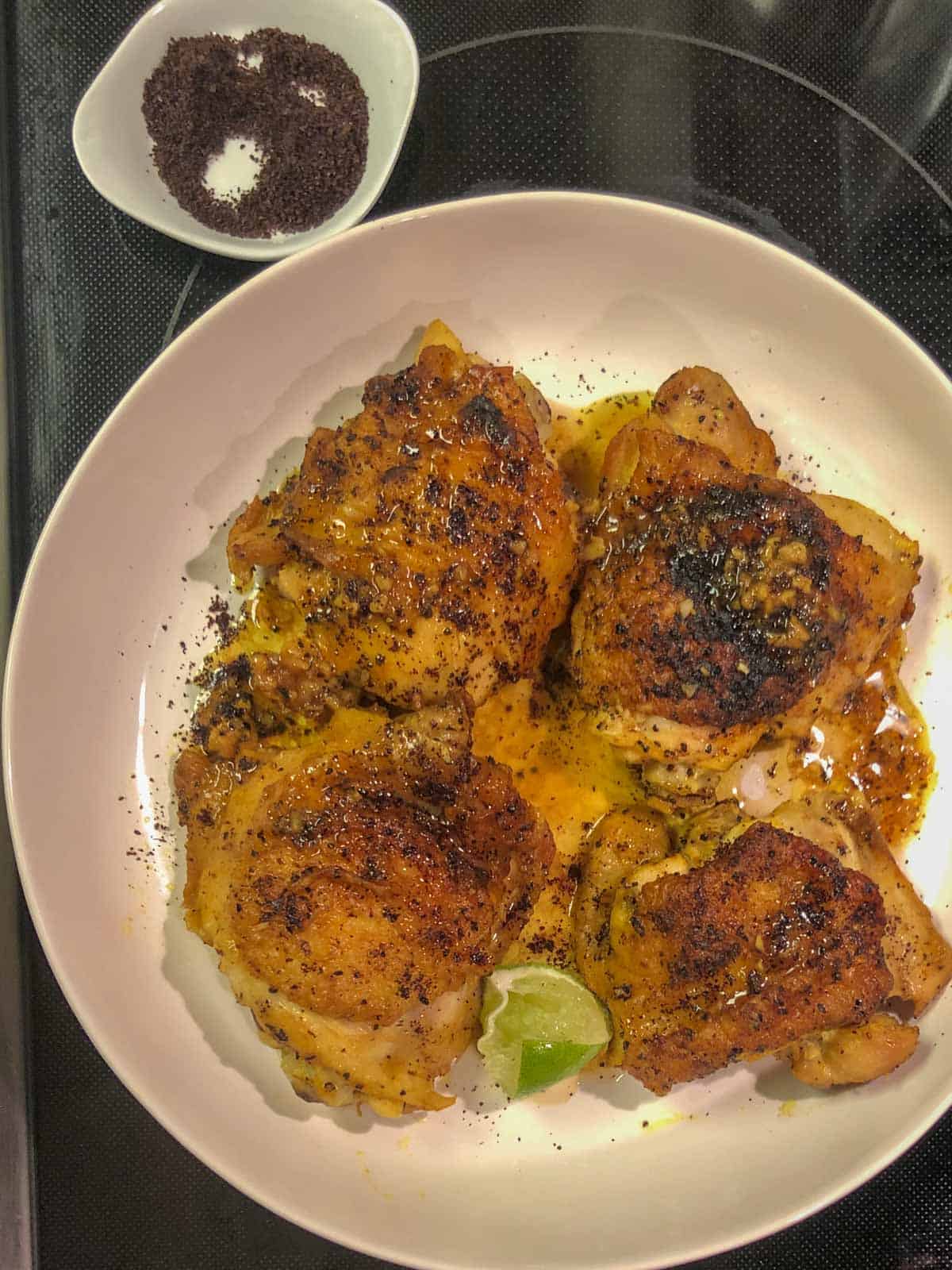 Browned and cooked chicken thighs sprinkled with sumac and sitting in a yellow tinged braising sauce with lime wedges in a white dish with some sumac in a small white dish in the background.
