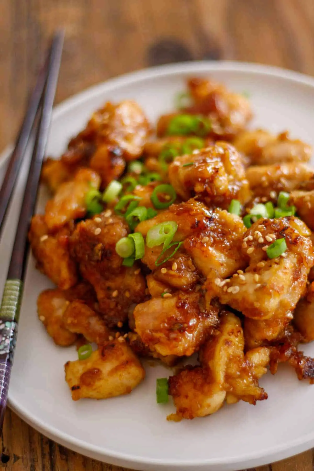 Chicken pieces with a sticky sauce on a white plate garnished with green onions and a pair of chopsticks resting on the side of the plate.