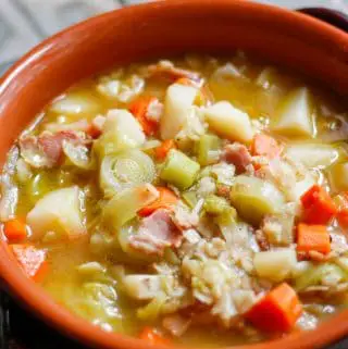 A terracotta soup bowl full of soup consisting of chicken broth, bacon, leeks, carrots, cabbage and potatoes.