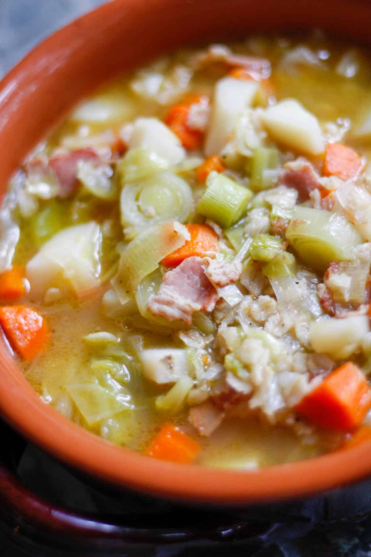 A terracotta soup bowl full of soup consisting of chicken broth, bacon, leeks, carrots, cabbage and potatoes.