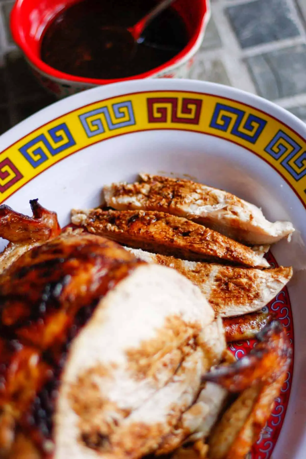 Sliced pieces of cooked chicken and a portion of a whole chicken next to the slices on a patterned plate with a red bowl filled with a soy based sauce next to the plate some of the sauce has been drizzled over the chicken.