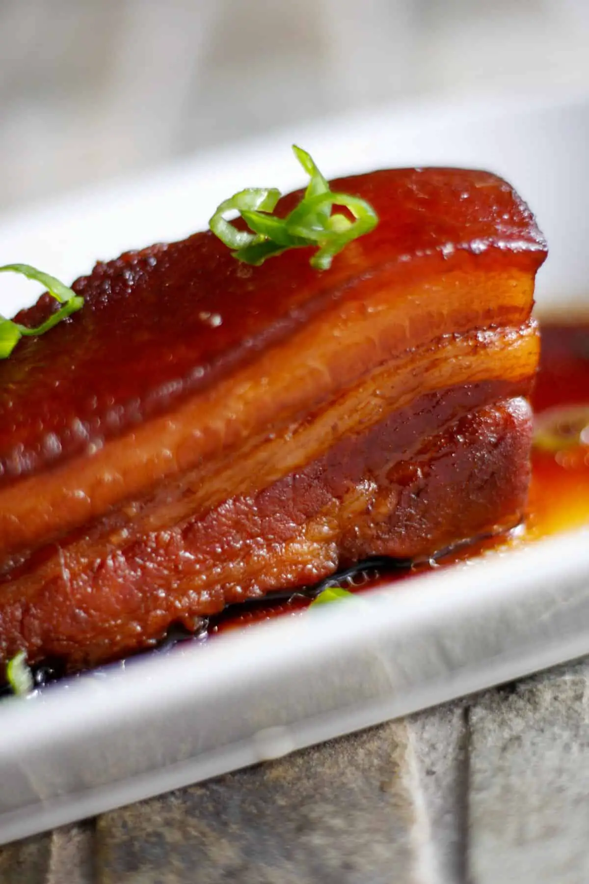 Braised pork belly that is reddish in color on a white dish atop braising sauce and garnished with green onions.