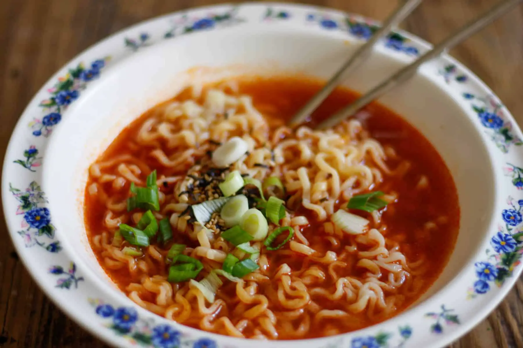 Ramen noodles in a spicy sauce garnished with seaweed, sesame seeds, and green onions in a white bowl patterned with blue flowers and silver chopsticks resting on the bowl.