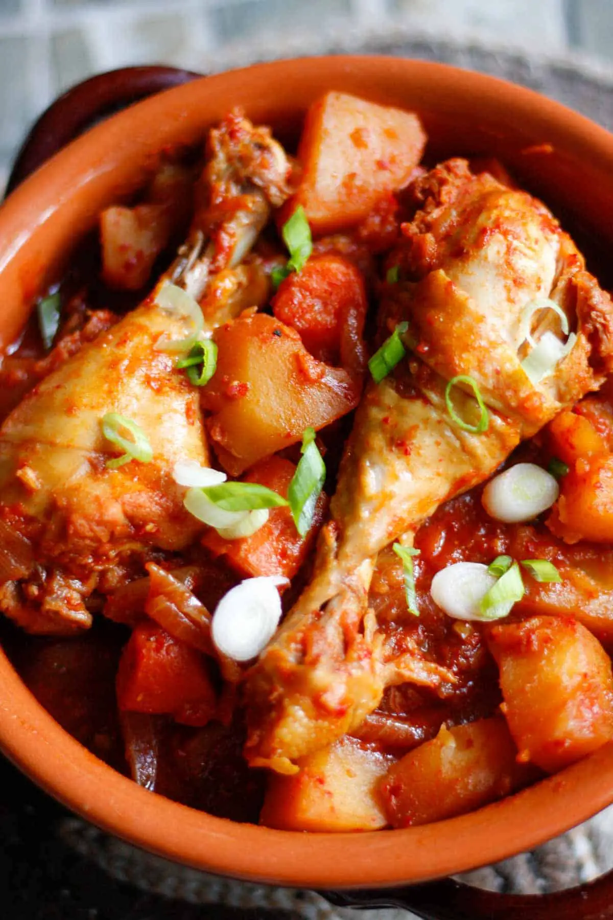 Chicken drumsticks in a spicy sauce with pieces of potato and carrots garnished with green onions in a bowl.