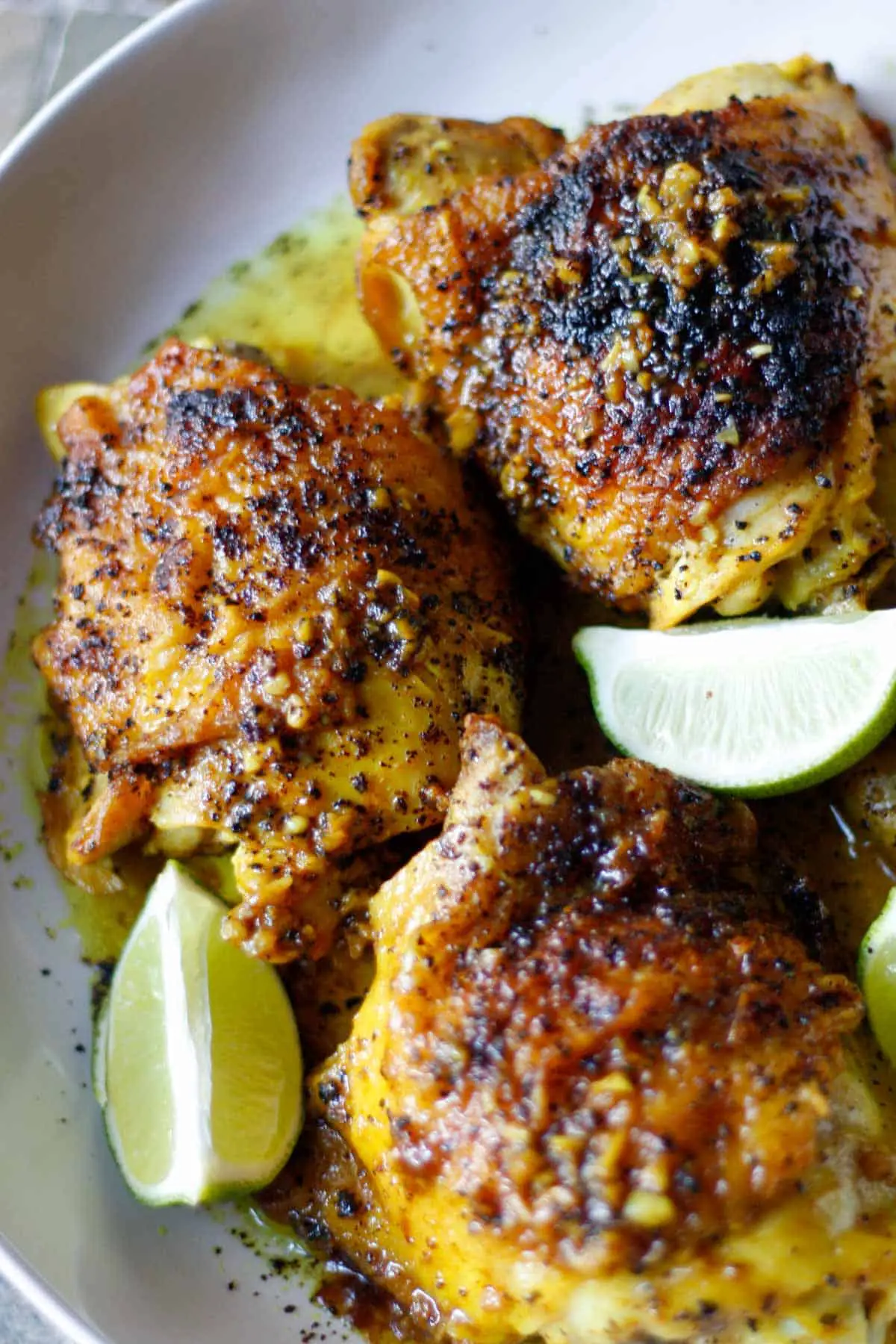 Browned and cooked chicken thighs sprinkled with sumac and sitting in a yellow tinged braising sauce with lime wedges in a white dish.