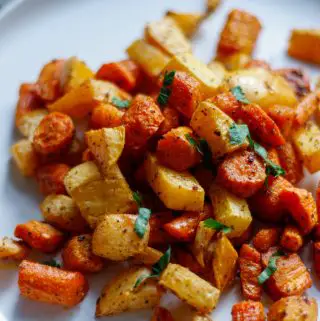 Pieces of carrots and rutabaga which have been roasted and placed on a white plate garnished with Italian parsley.