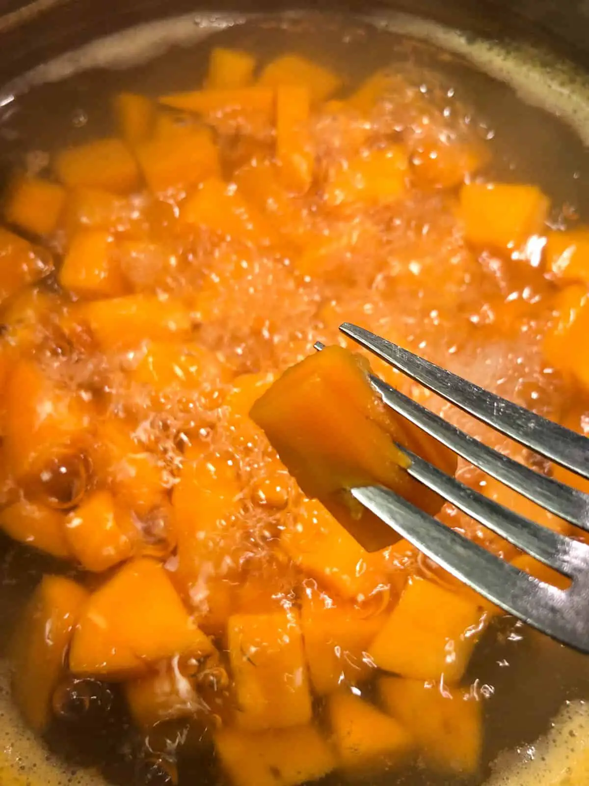 Cubes of sweet potatoes in boiling water in a saucepan with a fork piercing one of the sweet potato cubes in the foreground.