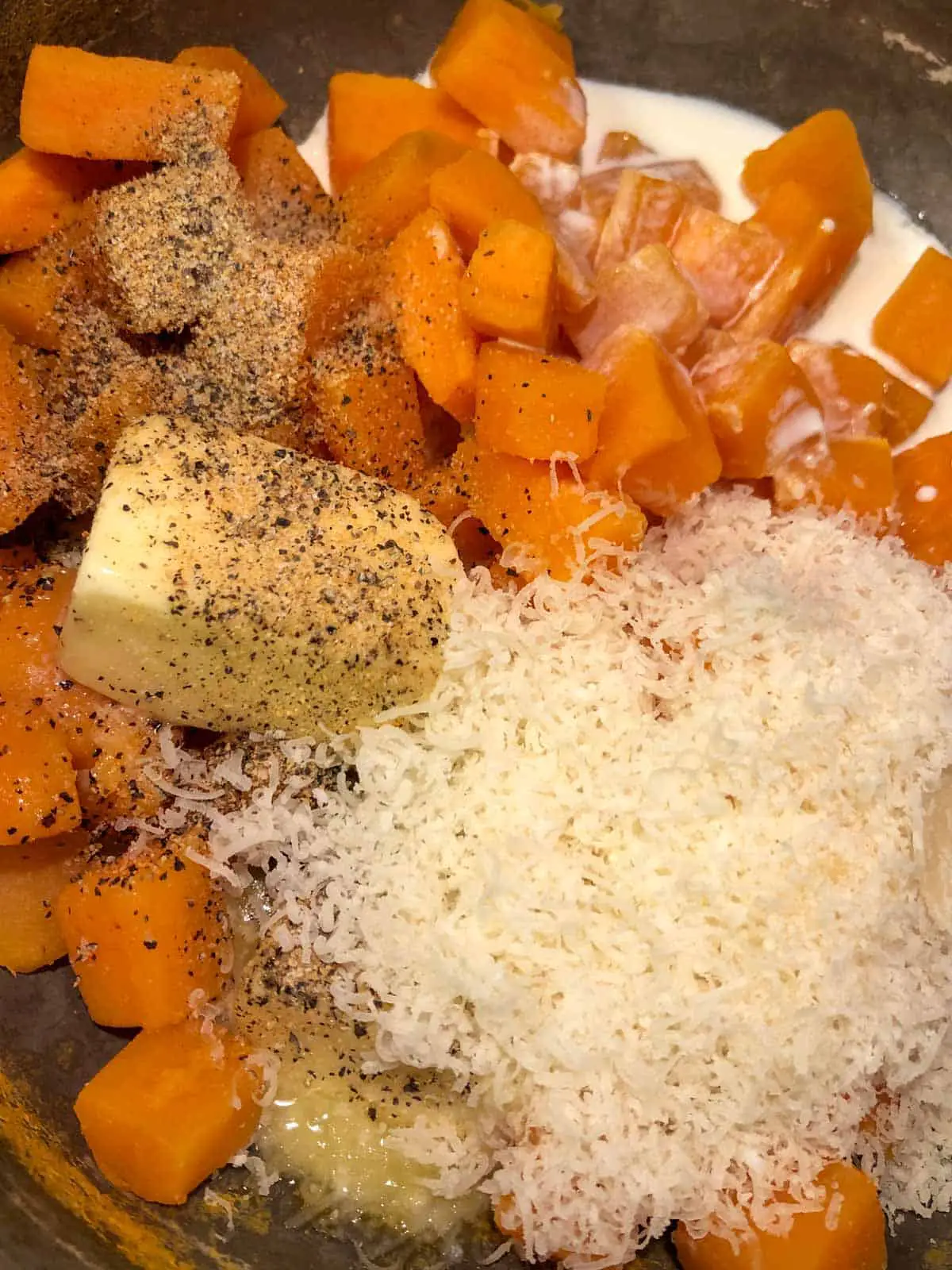 Cubes of sweet potatoes with cream, grated Parmesan cheese, butter, and seasonings in a saucepan.