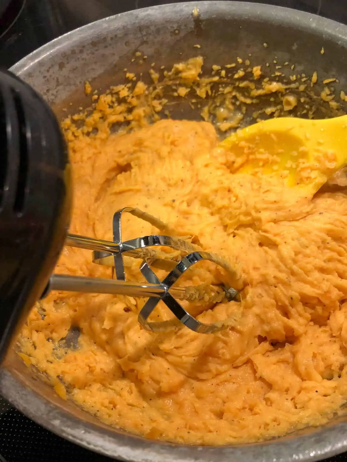Mashed sweet potatoes in a saucepan with a yellow silicone spoon and the whisk attachments of a hand blender in the bed of mashed sweet potatoes.