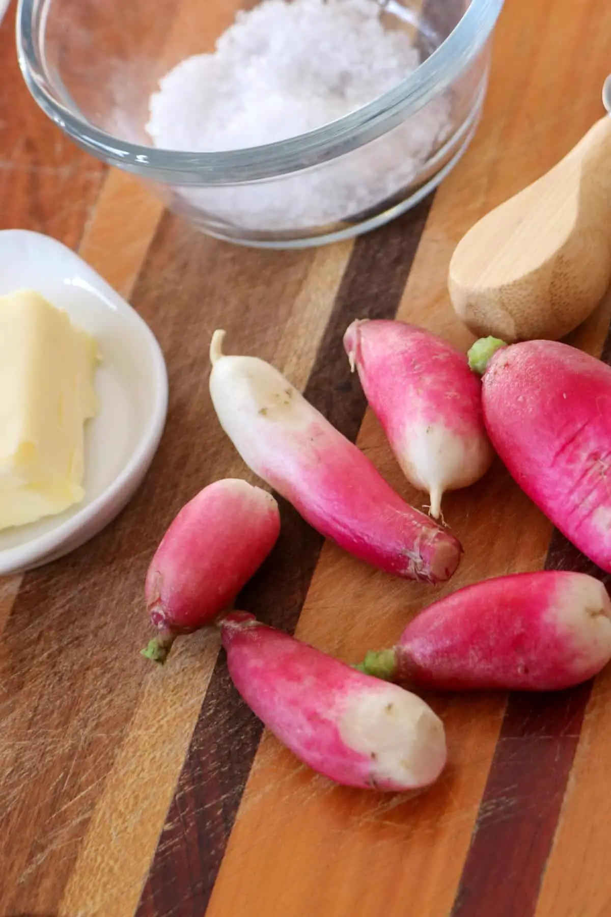 French breakfast radishes on a wooden cutting board, glass bowl with sea salt in the background, white dish with salt on the left, and top right a wooden handle of a cheese knife.