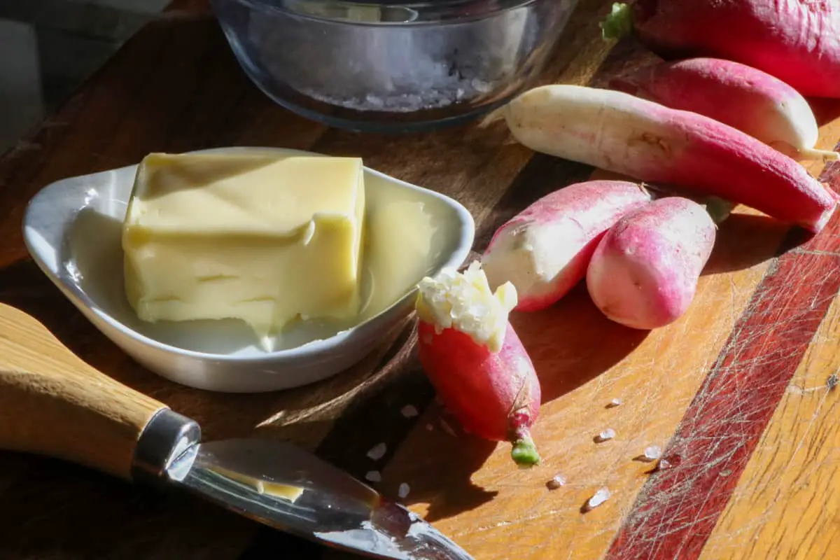 Several French breakfast radishes on a wooden cutting board with a glass bowl with sea salt in the background, a white dish with butter on the left side, and a cheese knife with wooden handle in the foreground.